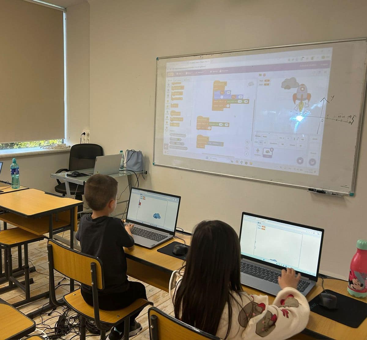 Doi elevi lucrează în Scratch la laptopuri, cu proiectul afișat pe tablă și axele x și y desenate lângă proiecție